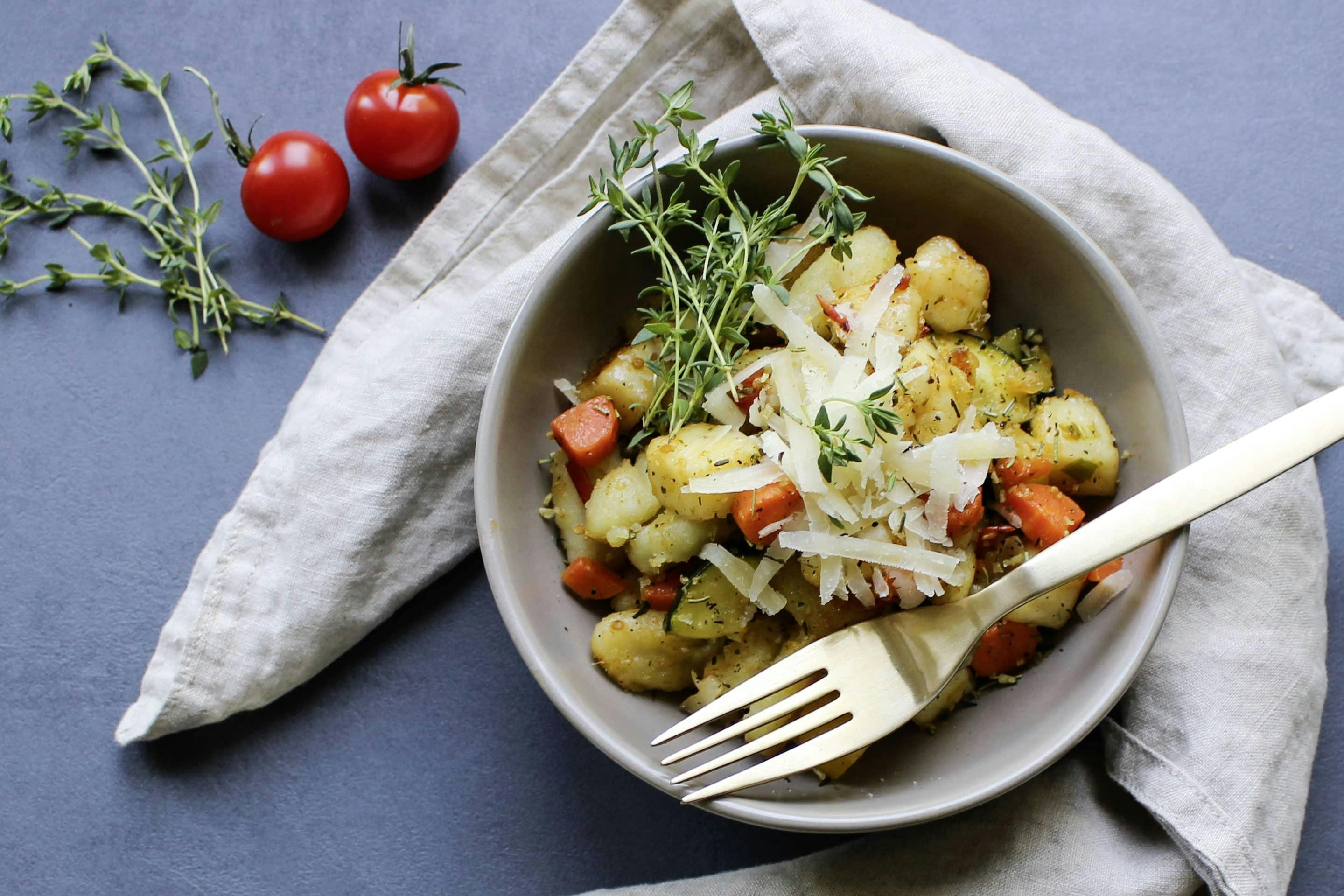 Gnocchi mit Grillgemüse und Halloumi in einer hellgrauen Schale mit goldener Gabel, Parmesan und frischen Thymianzweigen angerichtet