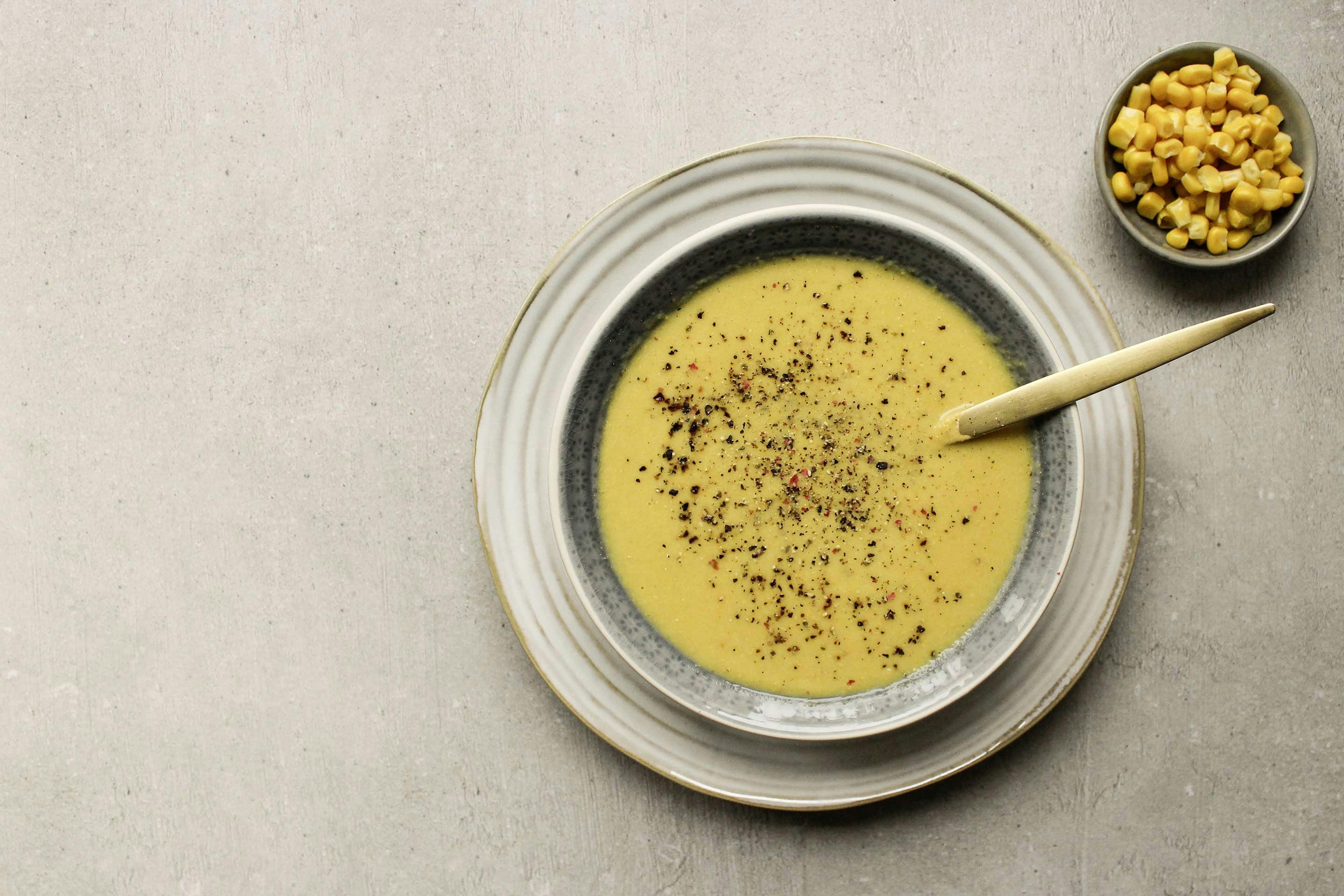 Maissuppe in einem tiefen Teller, mit Pfeffer garniert und einem goldenen Löffel angerichtet