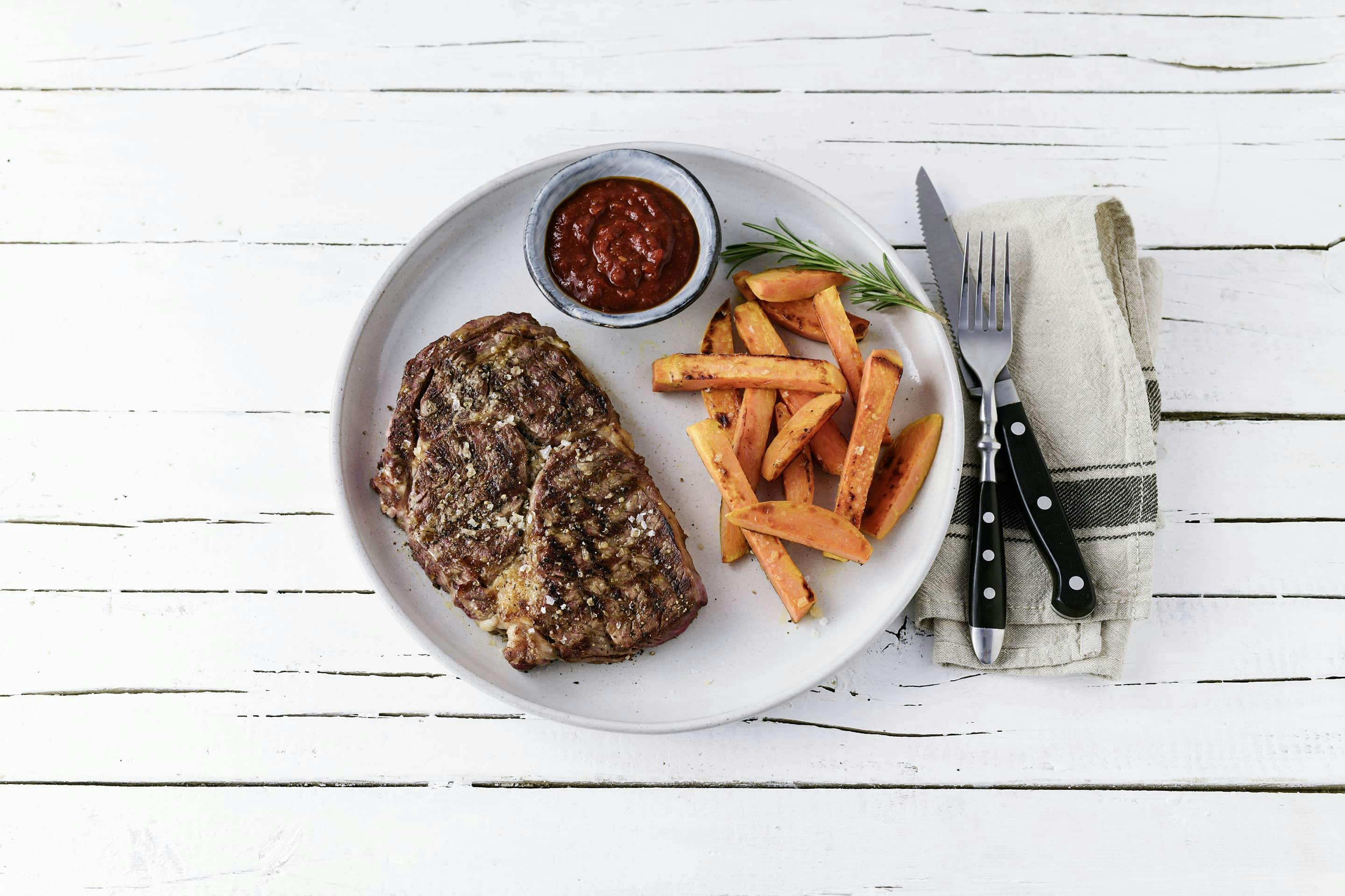 Magic-Dust-Steak mit Sueßkartoffelsticks und Sauce in einem Schälchen auf einem weißen Teller mit Besteck und Serviette
