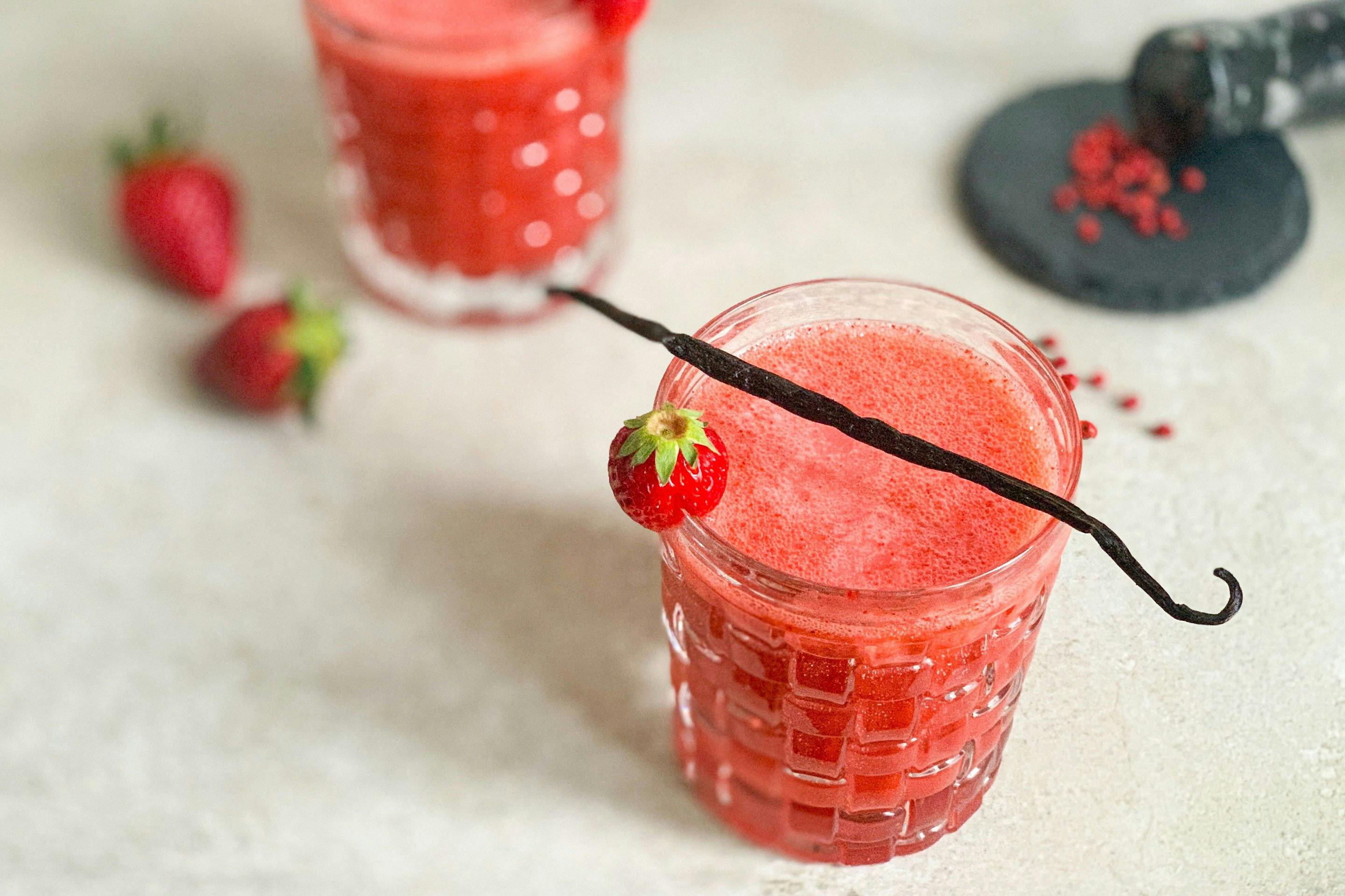 Kotányi Mule Cocktail in einem Glas mit Erdbeere und Vanilleschote garniert. Im Hintergrund befinden sich ein weiterer Cocktail und zerstoßene Rosa Beeren