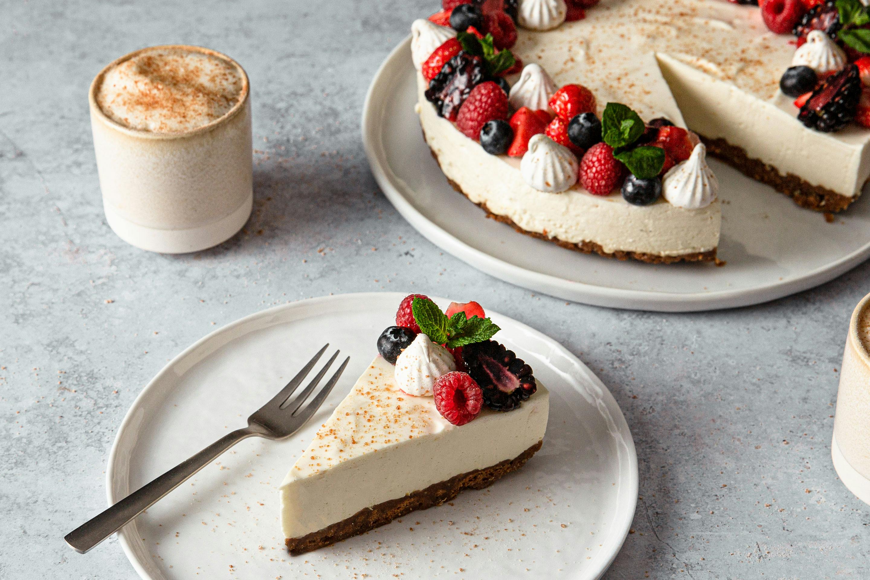 Cheesecake mit frischen Beeren garniert auf einer Kuchenplatte. Im Vordergrund ein Stück Cheesecake auf einem Teller mit Kuchengabel und einem Cappuccino in einem Becher.