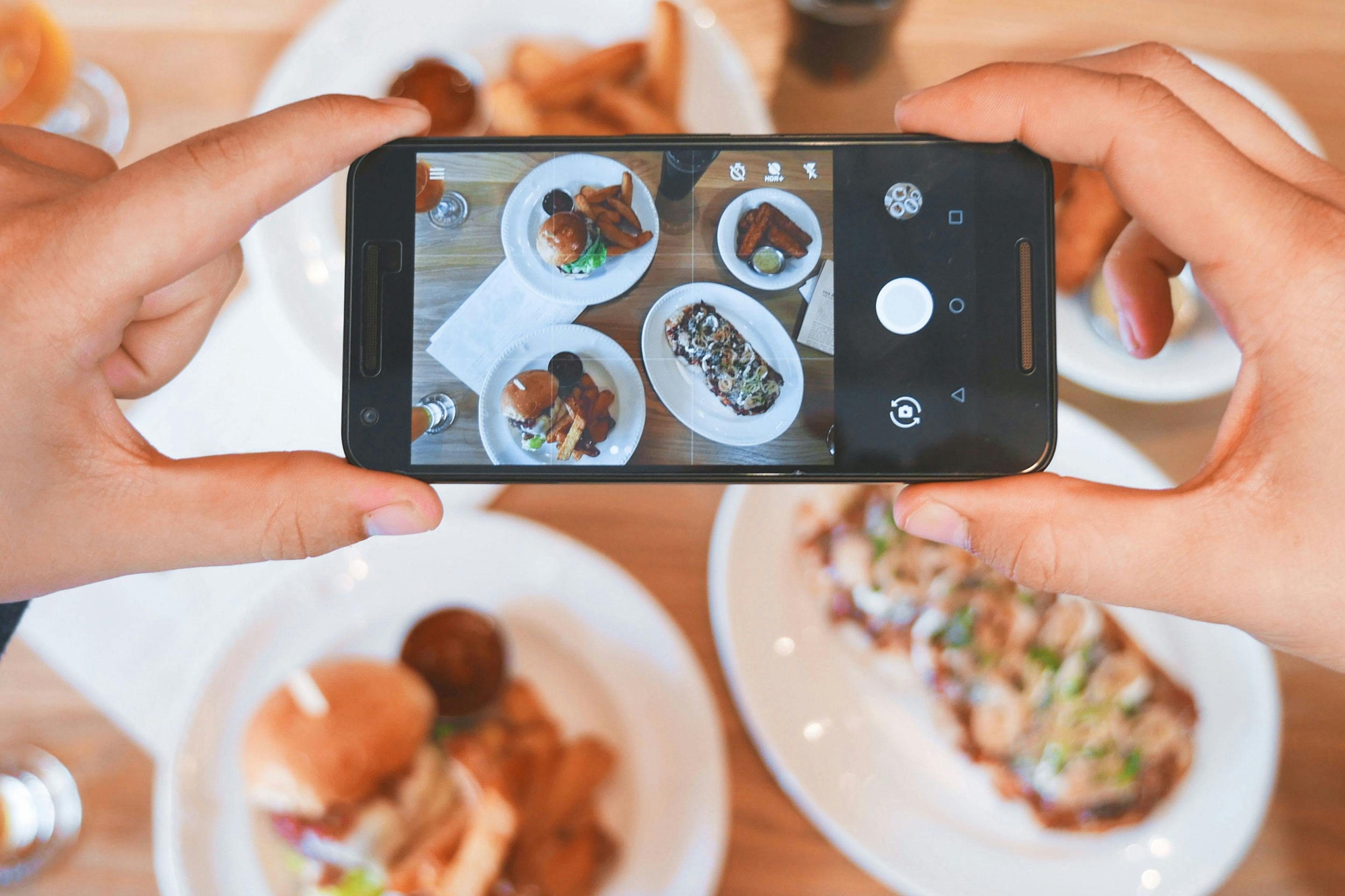 Männerhände halten ein Smartphone mit dem Fokus auf einen Tisch mit Essen gerichtet