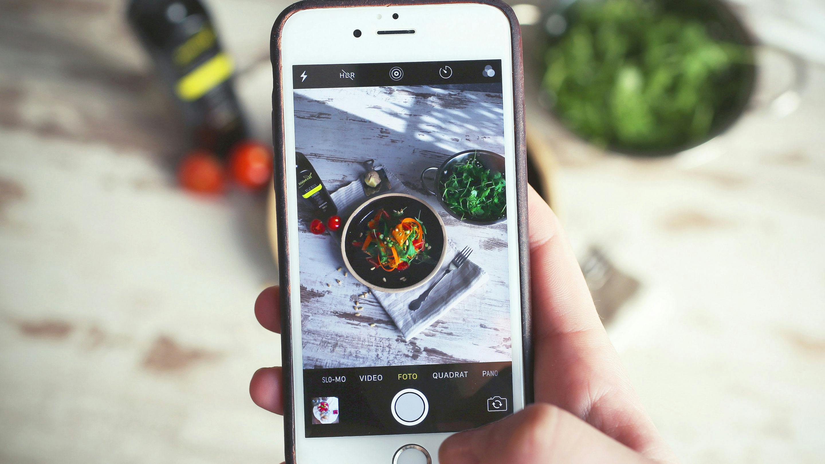 eine Hand hält ein Smartphone das gerade eine Holzplatte mit einem Teller voll frischem Salat fokussiert