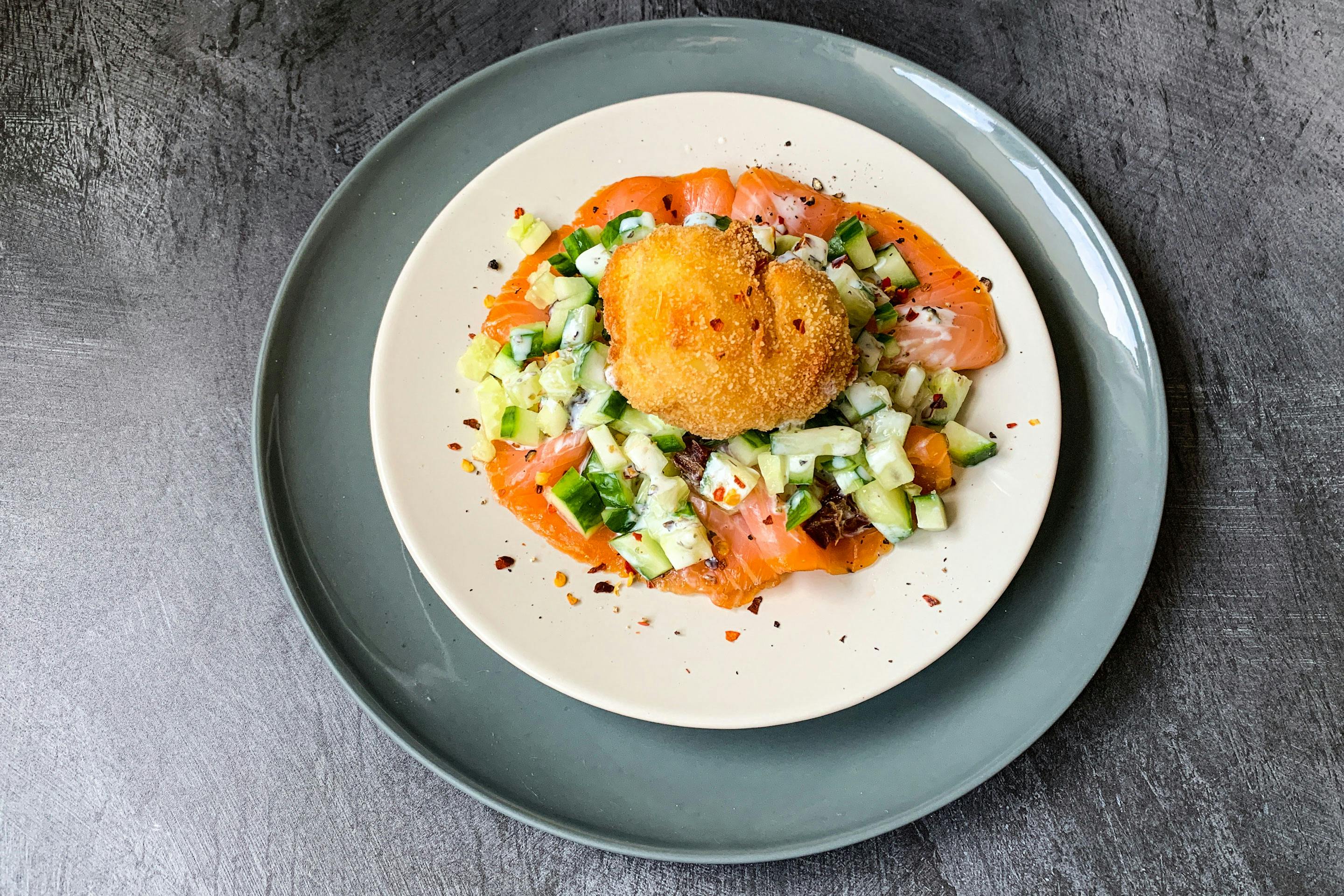 Dattel-Gurken-Salat mit Lachs und einem gebackenem Ei auf einem hellen Teller und einem grauen Platzteller