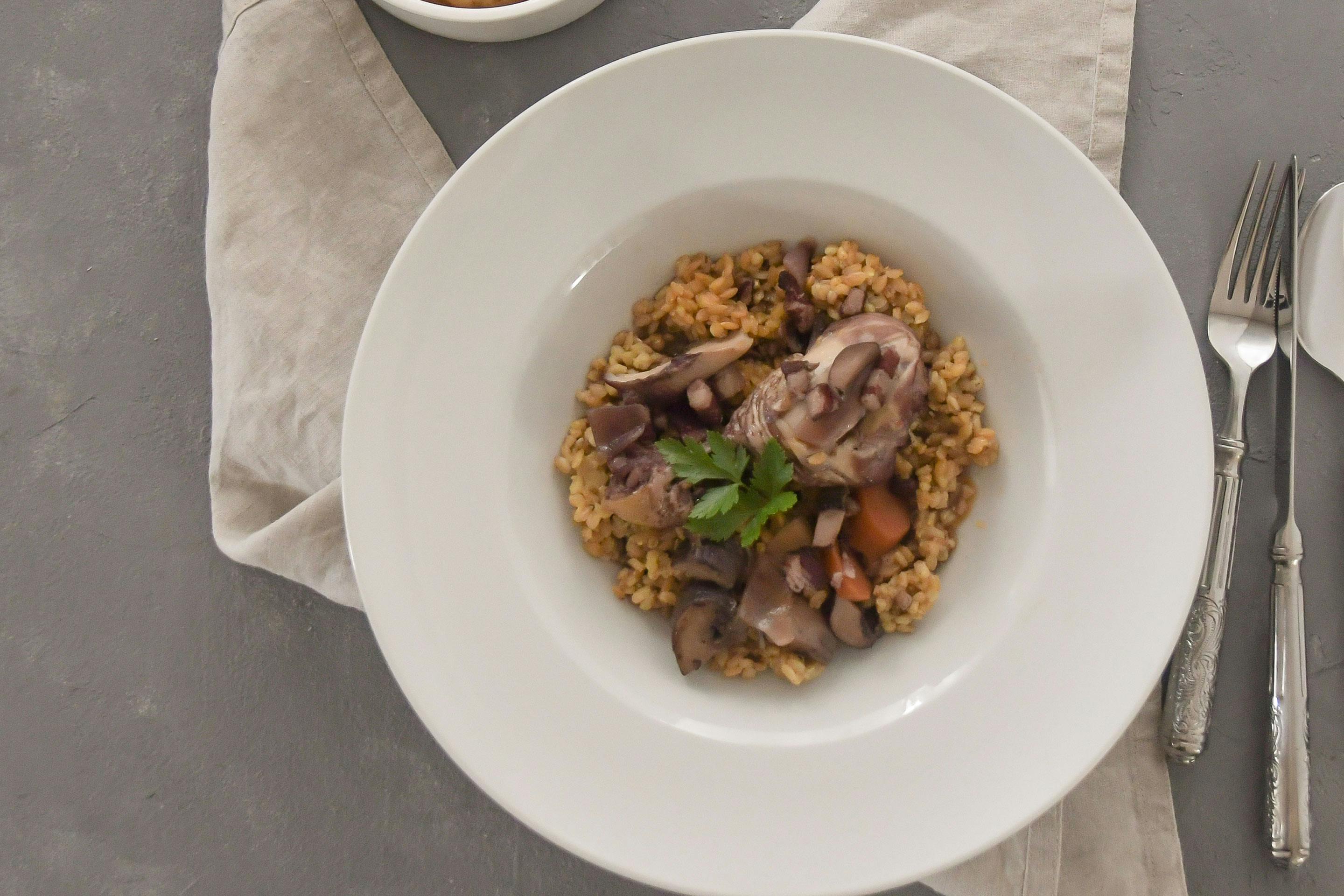 Coq Au Vin auf Einkornreis in einem tiefen Teller, dazu ein Schälchen mit Gemüse in Sauce auf grauem Untergrund