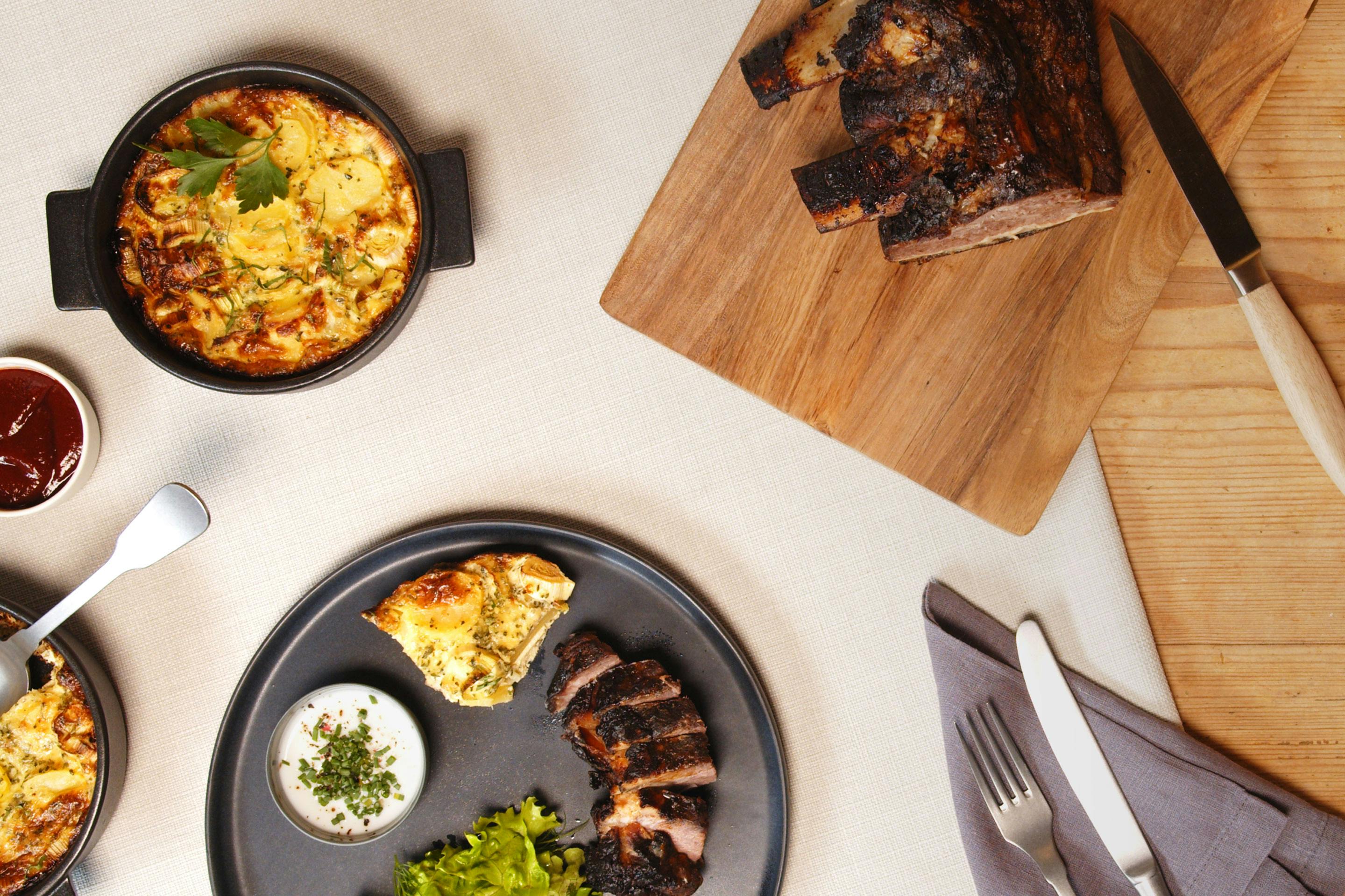 ein dunkler Teller mit aufgeschnittenen Beef Ribs und etwas Kartoffelgratin sowie Salatbeilage auf einer hellgrauen Tischdecke