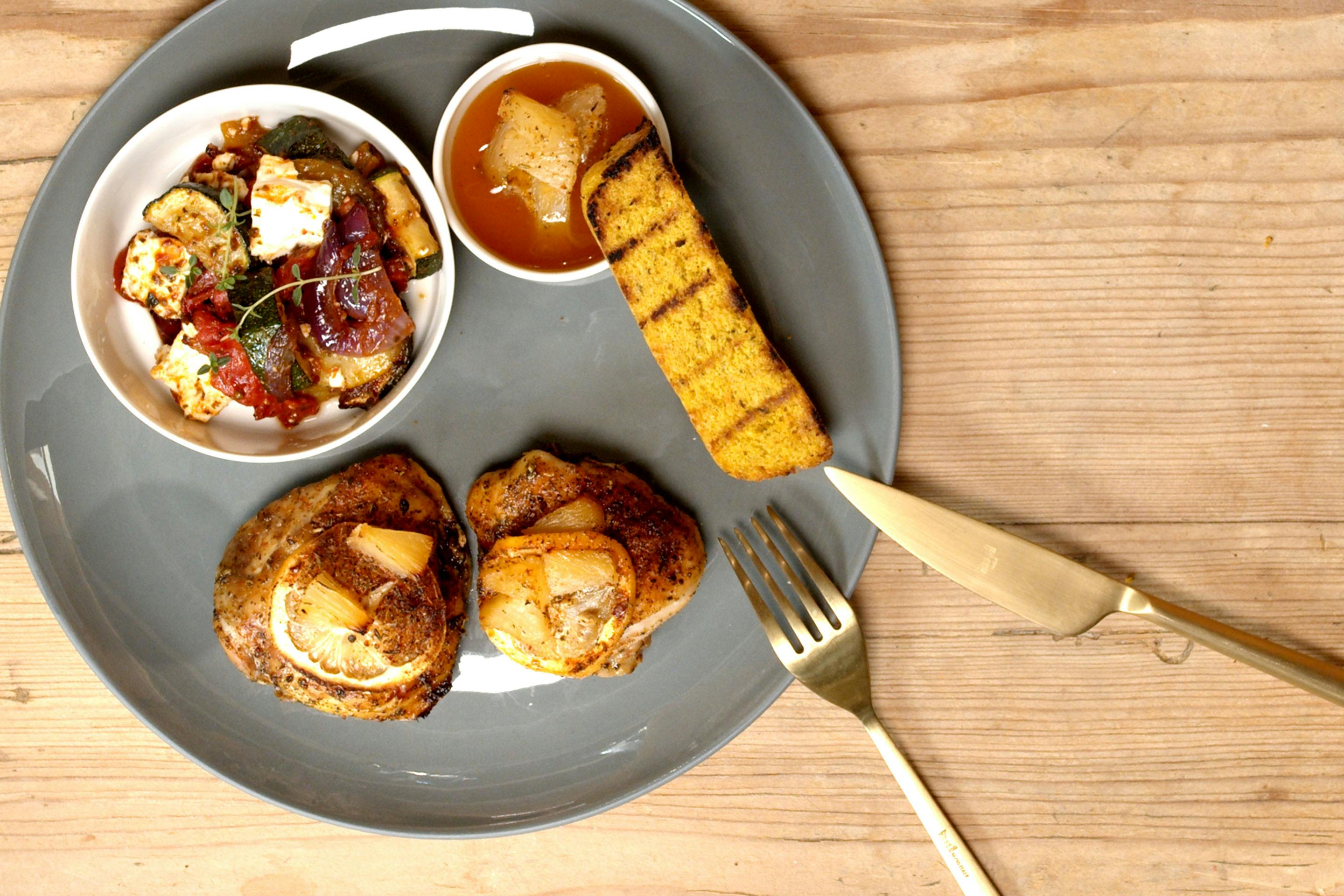 Huhn Hawaii Style mit gegrilltem Polentasterz auf einem dunkelgrauen Teller mit einem Schälchen voller Ratatouille, mit goldenem Besteck