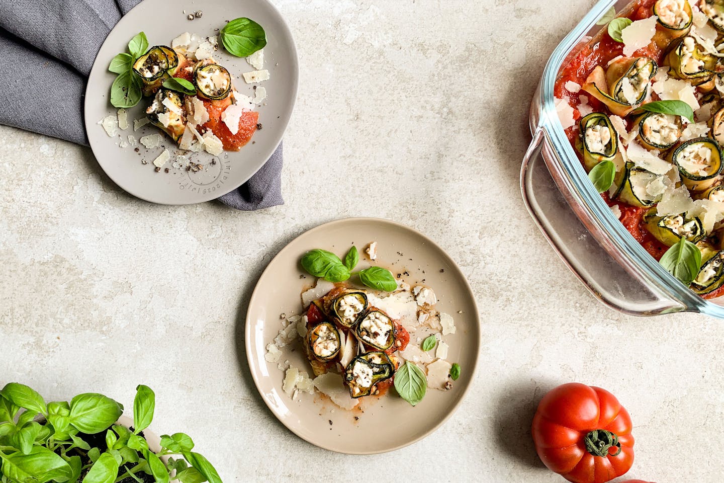 Zucchini-Canelloni auf 2 Tellern und in einer Auflaufform auf einem hellgrauen Untergrund, mit frischem Basilikum und einer Ochsenherztomate