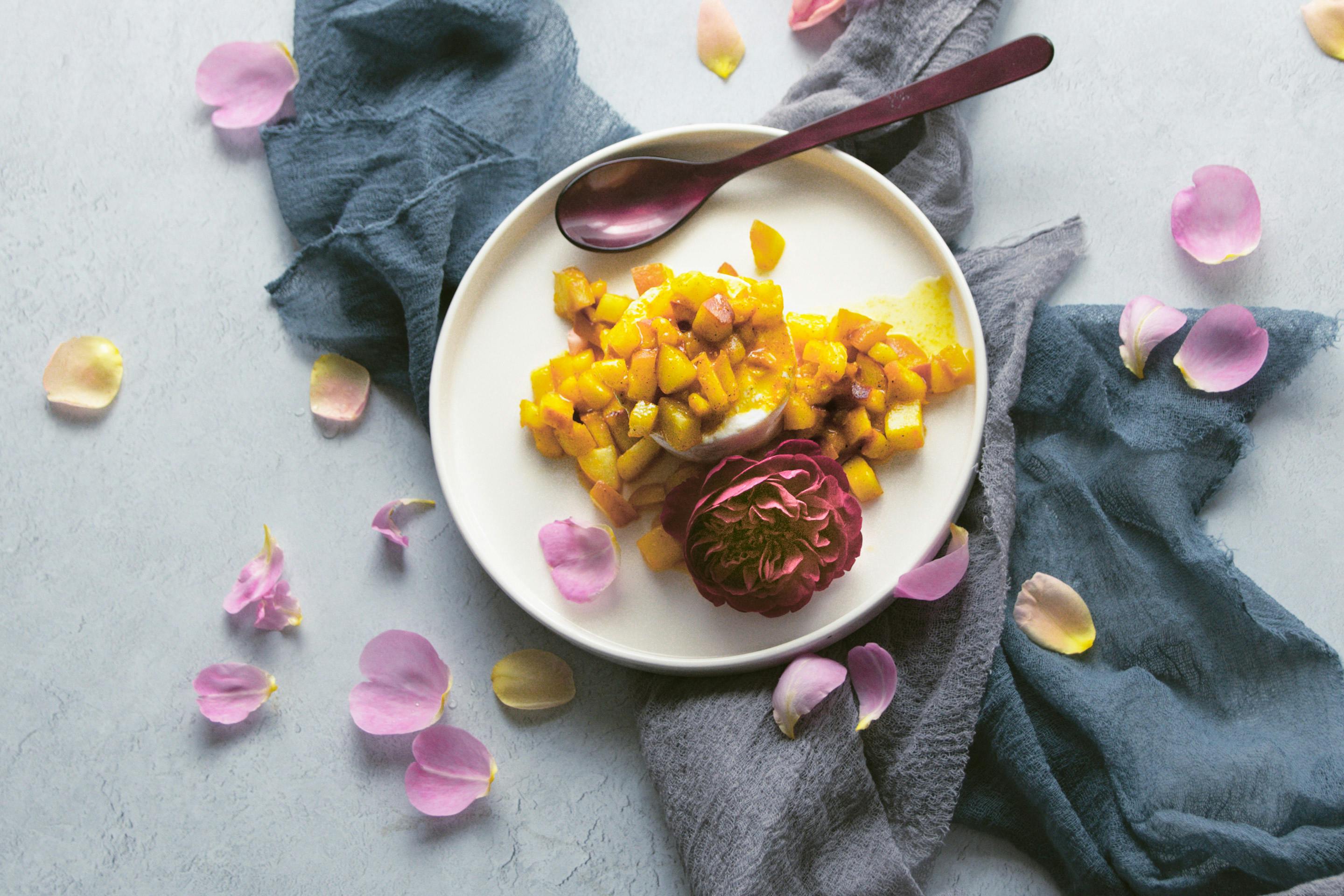 Rosenskyr mit fruchtigen Pfirsichstüchen und Vanillezucker mit Rosenblüte und -blättern auf einem hellen Teller angerichtet