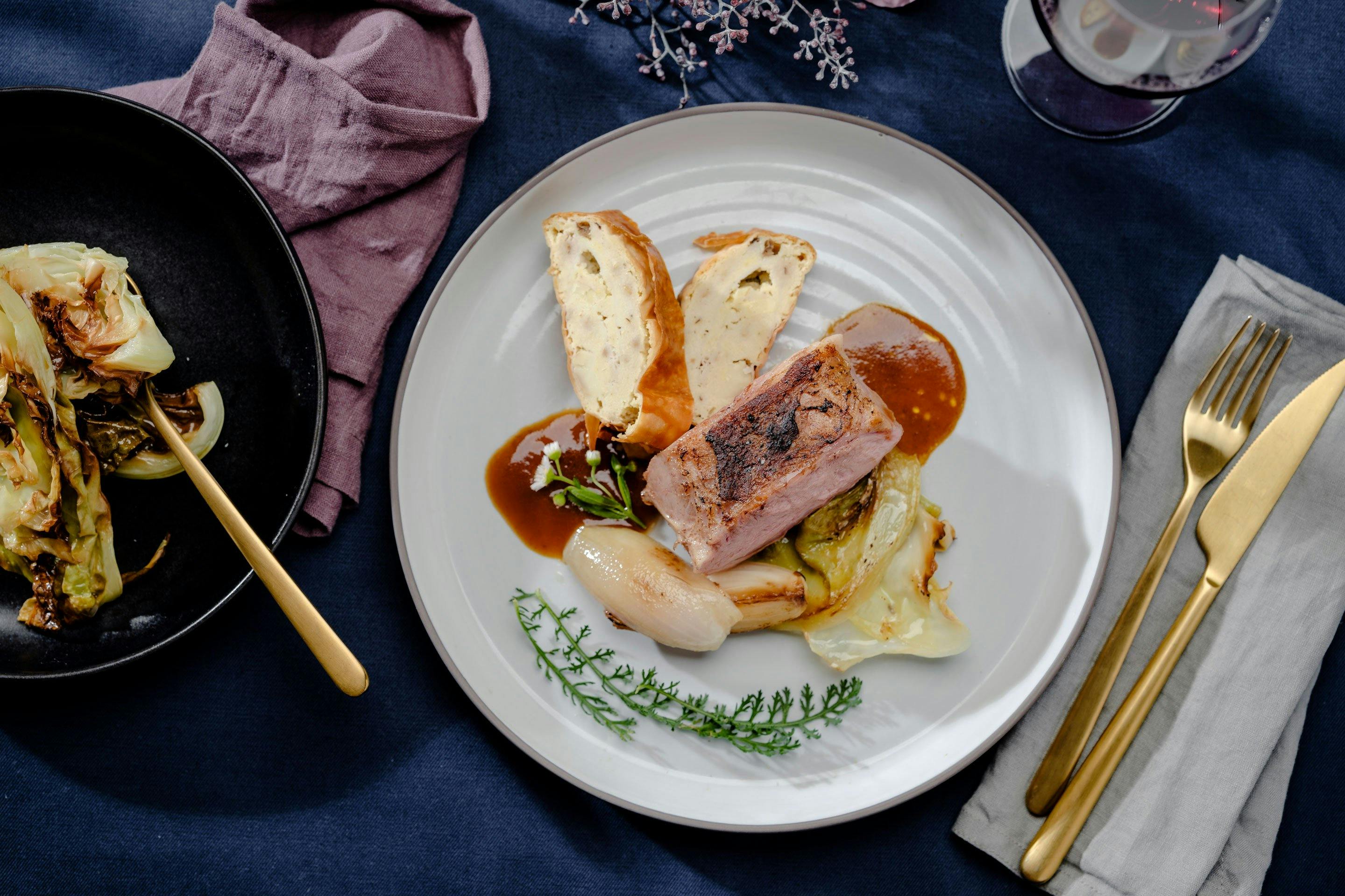 Kalbsbraten auf Ofenkraut mit Schalotten und Griessstrudel auf einem beigen Teller mit einem Glas Rotwein und daneben einem schwarzen Teller mit Kraut aus dem Ofen