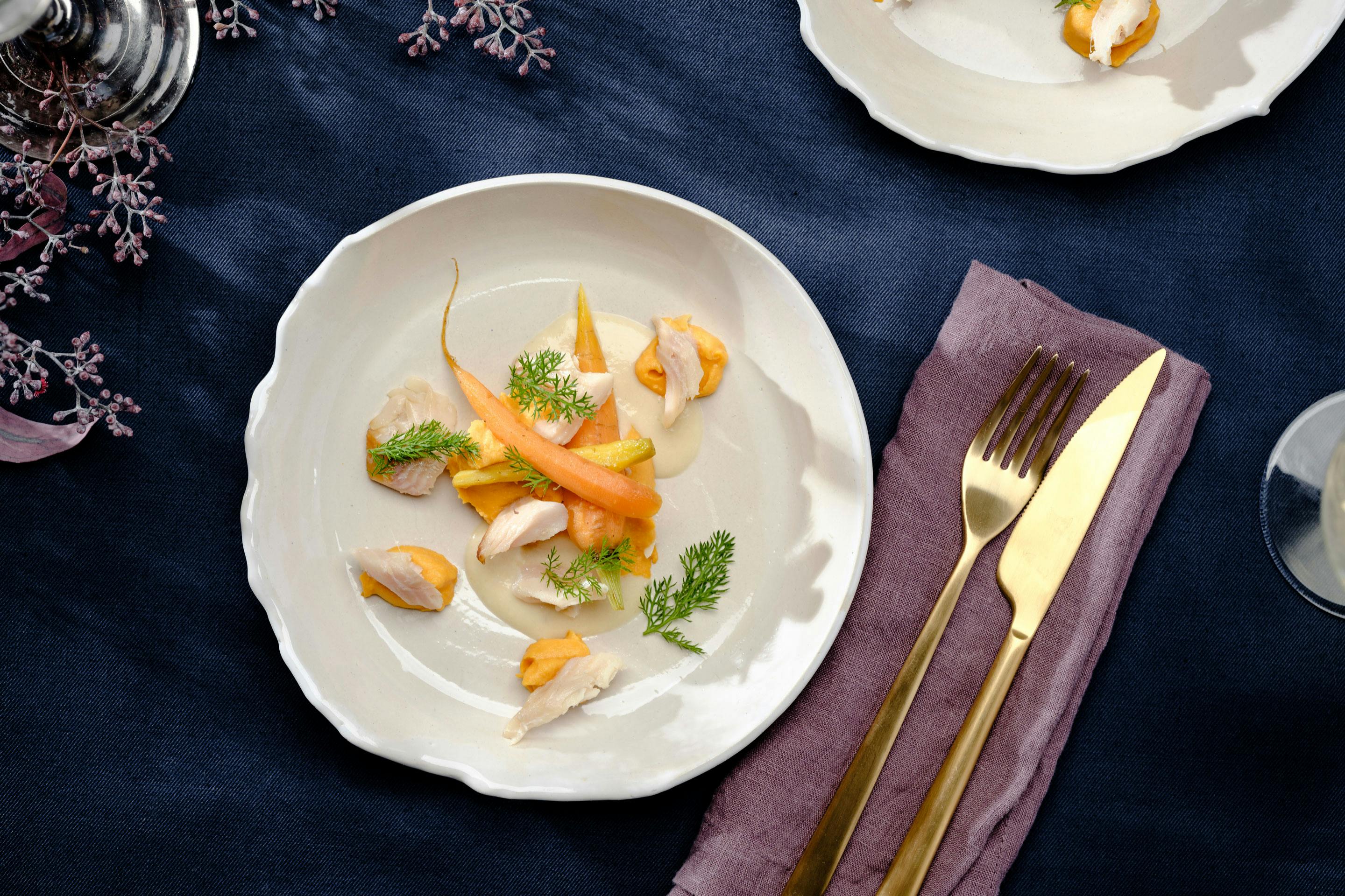 Zweierlei der Feldkarotte mit Karottenpürree und Räucherforellefiletstücken auf einem creme-farbenen Teller angerichtet mit goldenem Besteck auf dunkelblauer Tischdecke