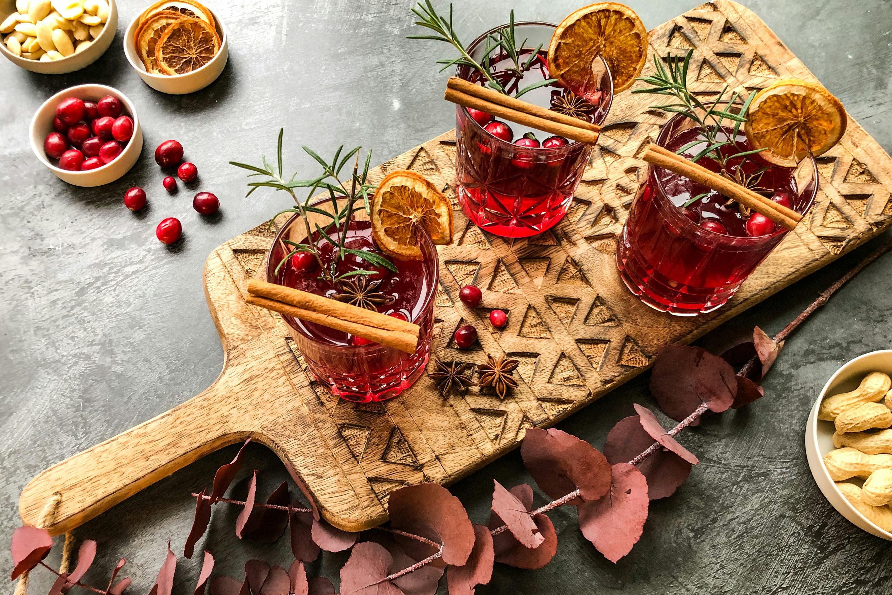 3 Gläser mit Cranberry Spice Mimosas auf einem Holzbrett winterlich verziert mit Zimtstangen, Rosmarinzweigen und getrockneten Orangenscheiben