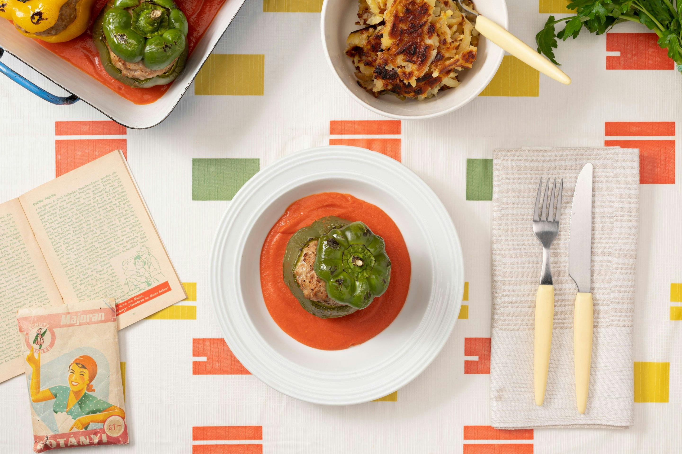 Bunte, gefüllte Paprika in einer Ofenform mit Tomatensauce, geröstete Kartoffeln als Beilage und ein weißer Teller mit einer grünen Paprika auf einem Tischtuch mit typischem fünfziger Jahre Muster.