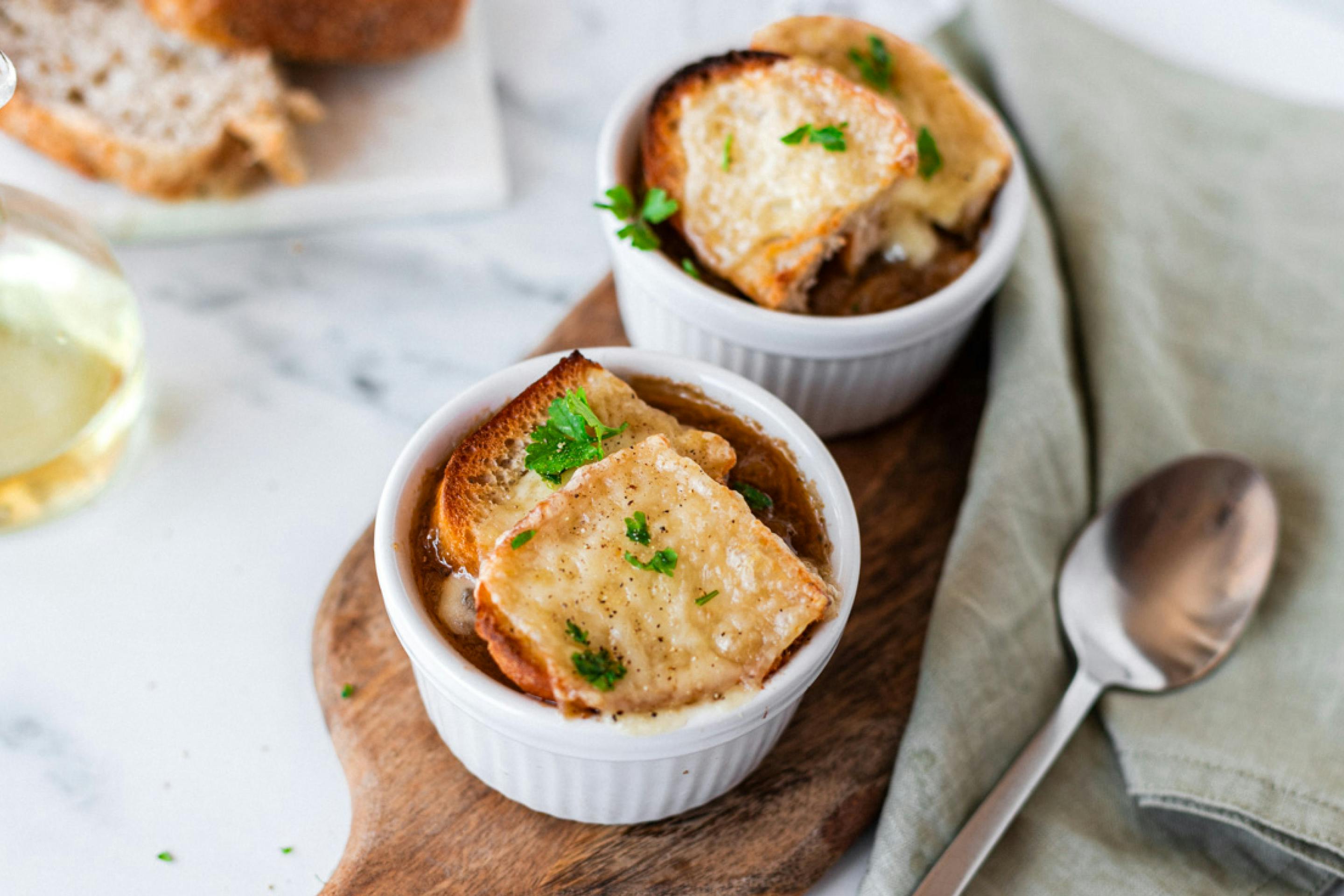 Two bowls of French onion soup topped with melted cheese and bread, garnished with parsley, served on a wooden board with a piece of bread and a knife on the side.