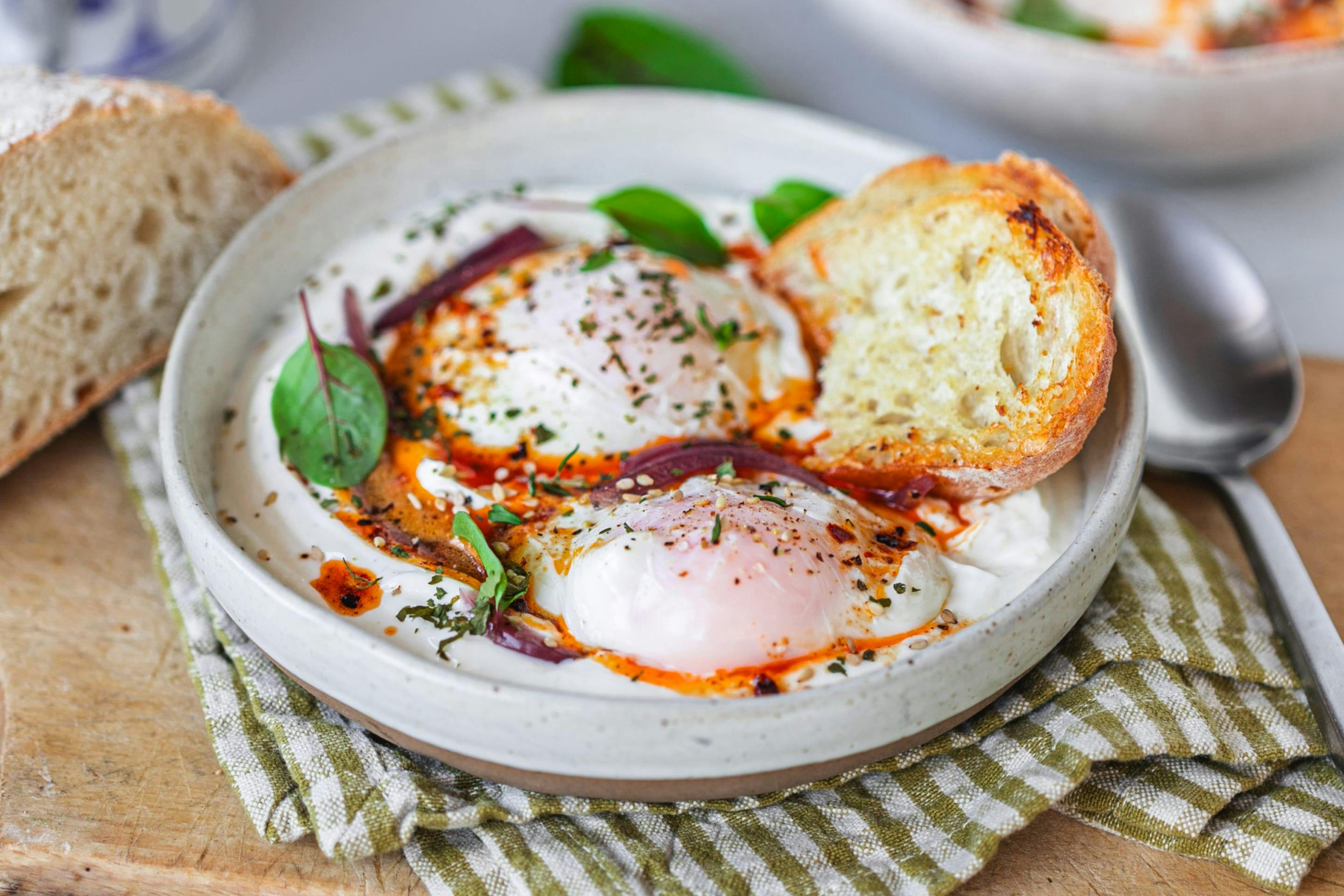 A dish featuring poached eggs served on yogurt with paprika butter, garnished with basil and red onions. Accompanied by slices of sourdough bread.